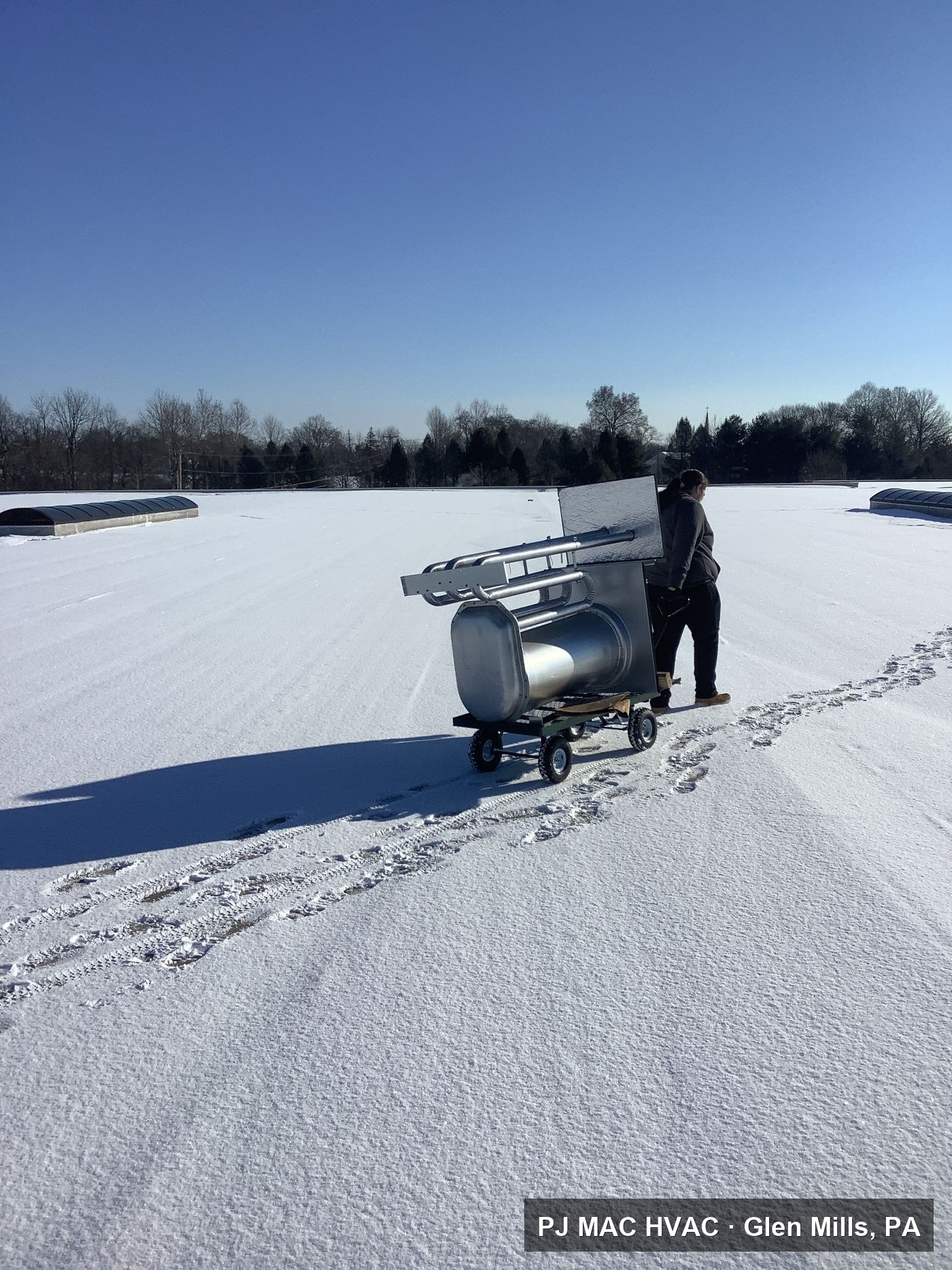A PJ MAC HVAC technician hauls replacement parts across a snow-covered commercial roof.